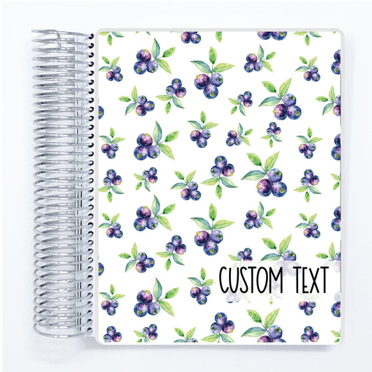 a spiral notebook with blueberries and leaves on a white background